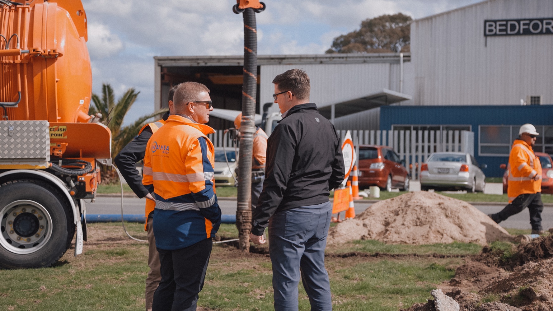 Two men, one wearing a hi-vis jacket with Waimea Group on the back, talking in front of an orange hydrovac being operated on a worksite.
