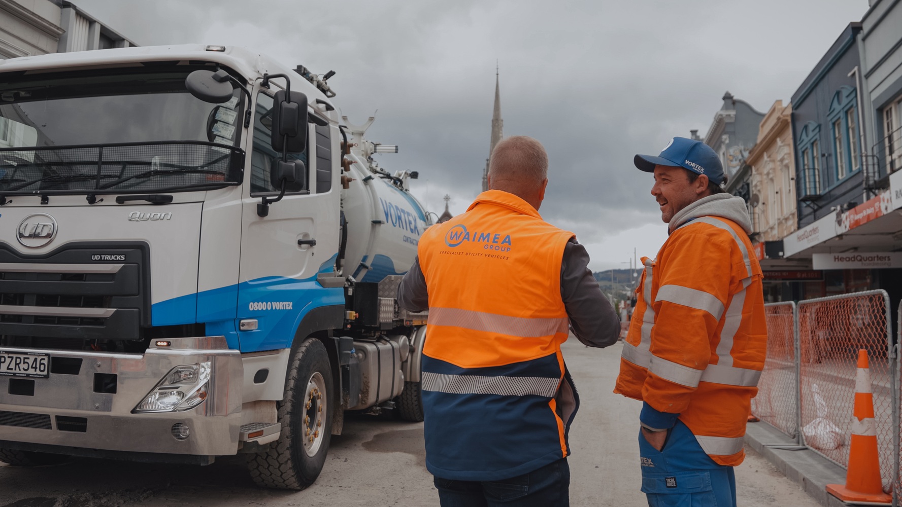 Two men, one wearing a hi-vis jacket with Waimea Group on the back, talking in front of an orange hydrovac being operated on a worksite.