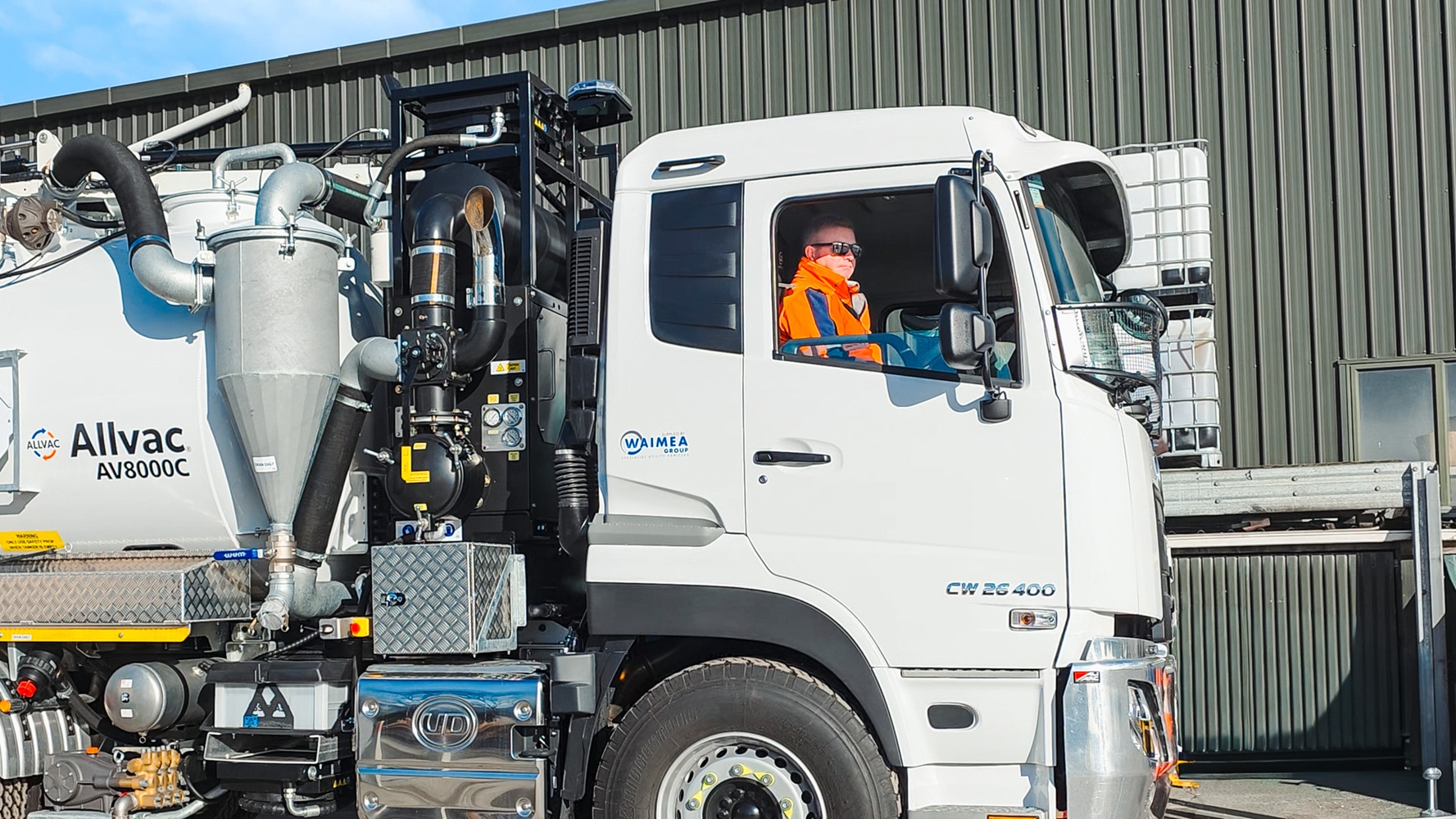 Two men, one wearing a hi-vis jacket with Waimea Group on the back, talking in front of an orange hydrovac being operated on a worksite.