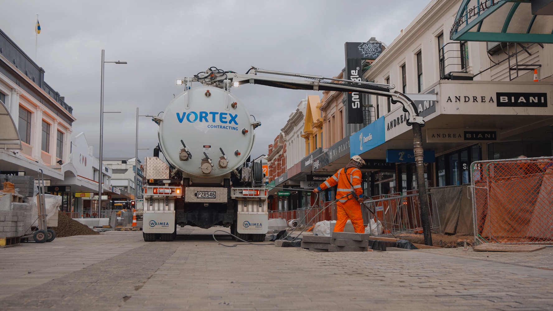 Two men, one wearing a hi-vis jacket with Waimea Group on the back, talking in front of an orange hydrovac being operated on a worksite.
