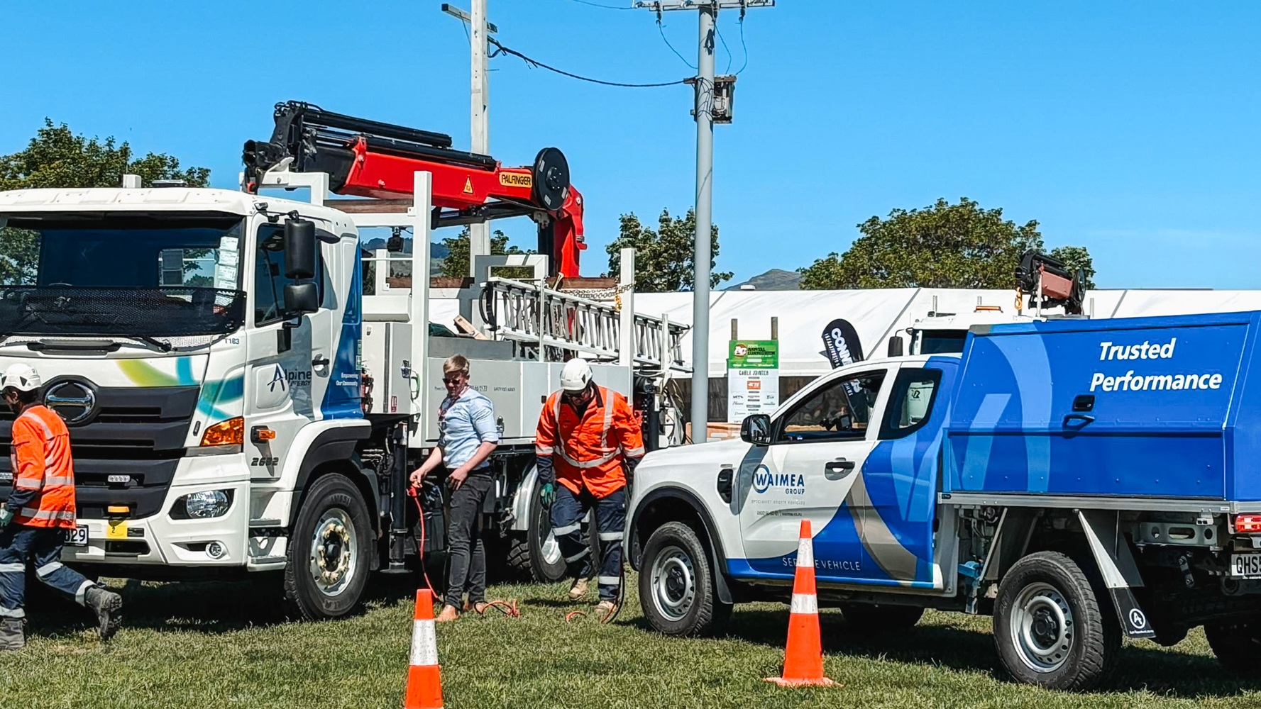 A showground viewed from up high, with lots of lines crane trucks set up with their crews demonstrating next to power poles, and stalls around the edge of the grounds, early in the morning.