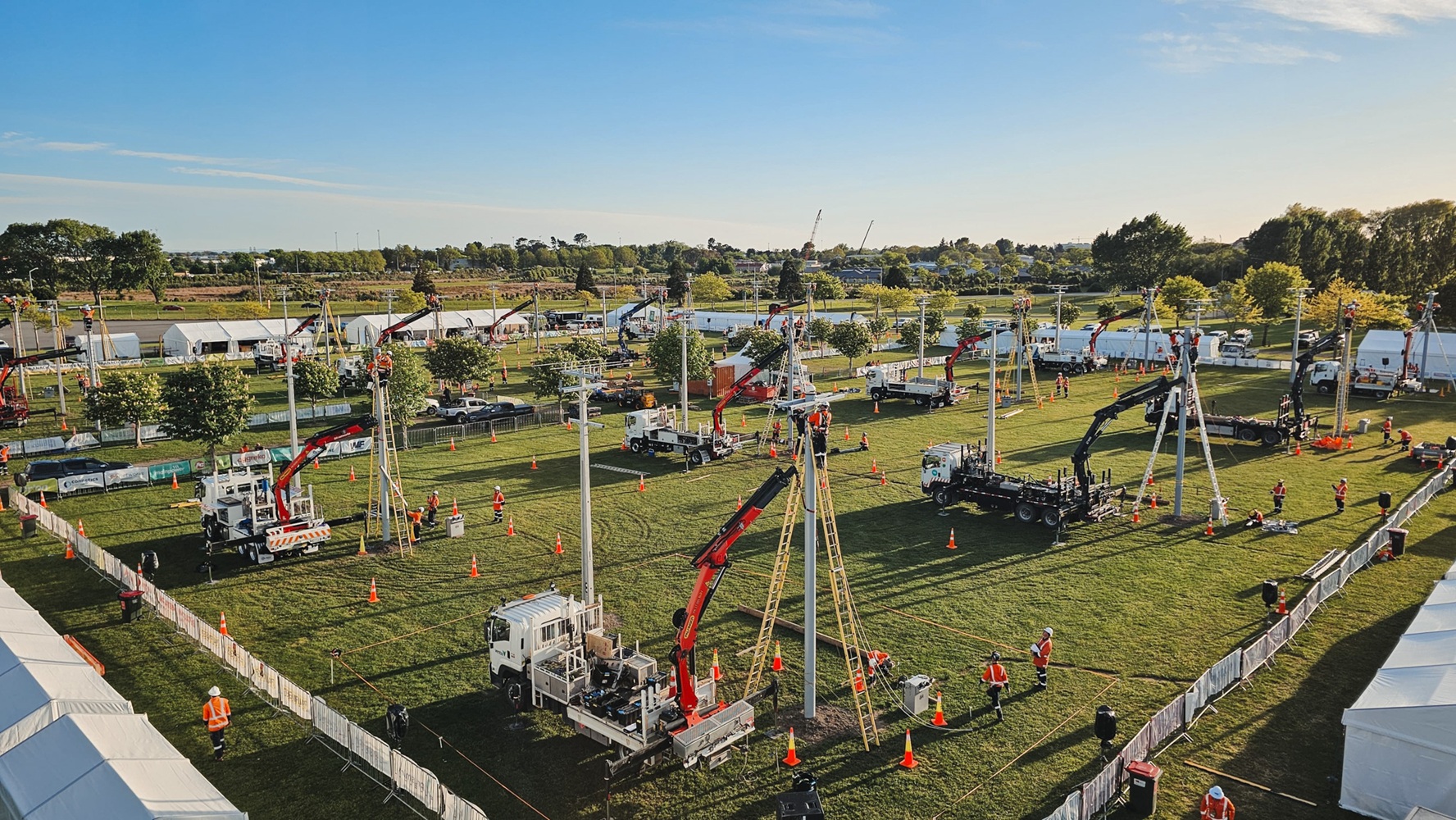A showground viewed from up high, with lots of lines crane trucks set up with their crews demonstrating next to power poles, and stalls around the edge of the grounds, early in the morning.